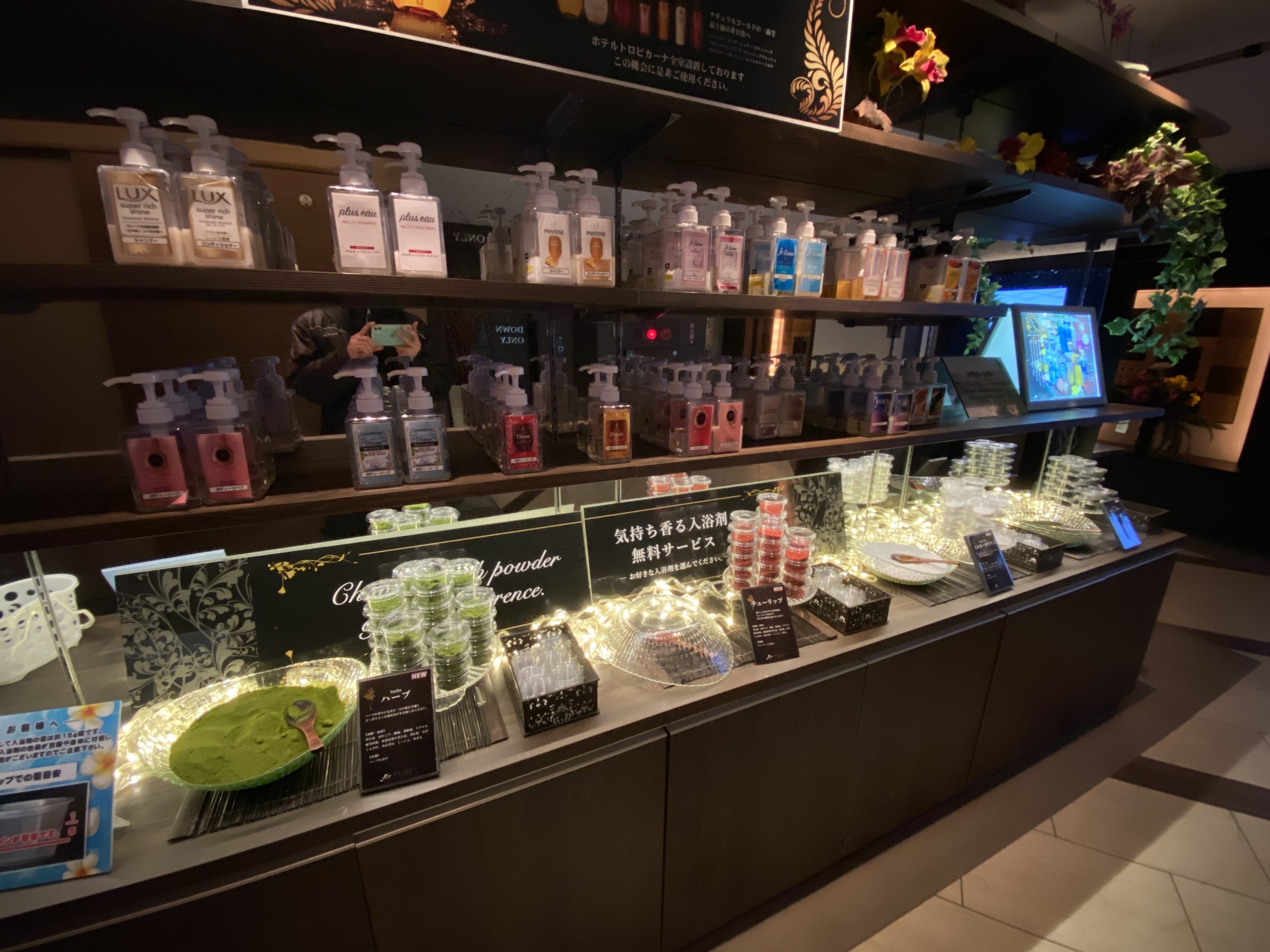 ホテルトロピカーナのエントランスに設置されたアメニティコーナー。棚には数種類のブランドシャンプーが並び、手前のカウンターには無料サービスの入浴剤が電飾と共に華やかにディスプレイされている